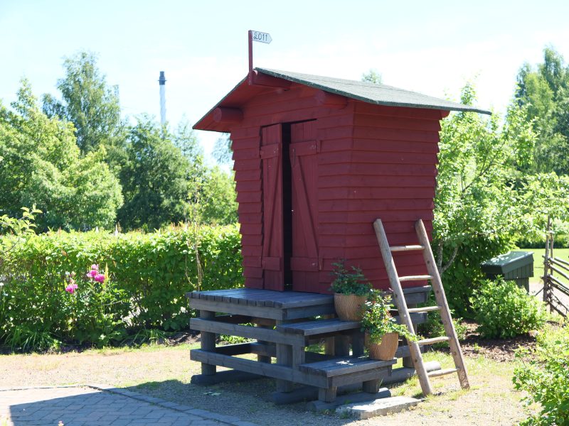 Punainen maitolaituri, harmaat portaat ja ja portailla kaksi kukkaruukkua kukkineen.